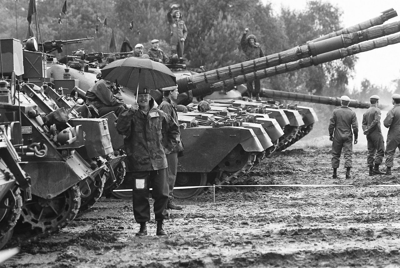 A soldier smiles while holding an umbrella and standing in the muddy tank ruts during exercise REFORGER 85 in Germany.