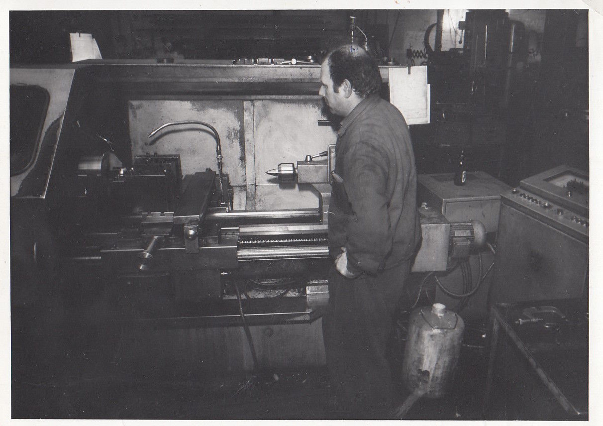 Vintage black-and-white photograph of a factory worker operating a large industrial lathe machine inside a workshop, showing hands-on manufacturing work, heavy equipment, and an industrial production environment