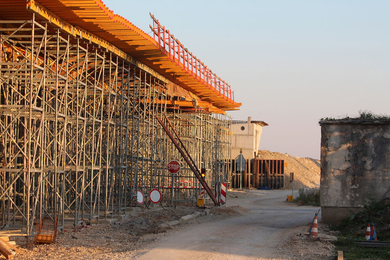 A large-scale road or bridge is under construction, featuring dense metal scaffolding, overhead formwork, stop signs, and gravel roadways marking restricted site access