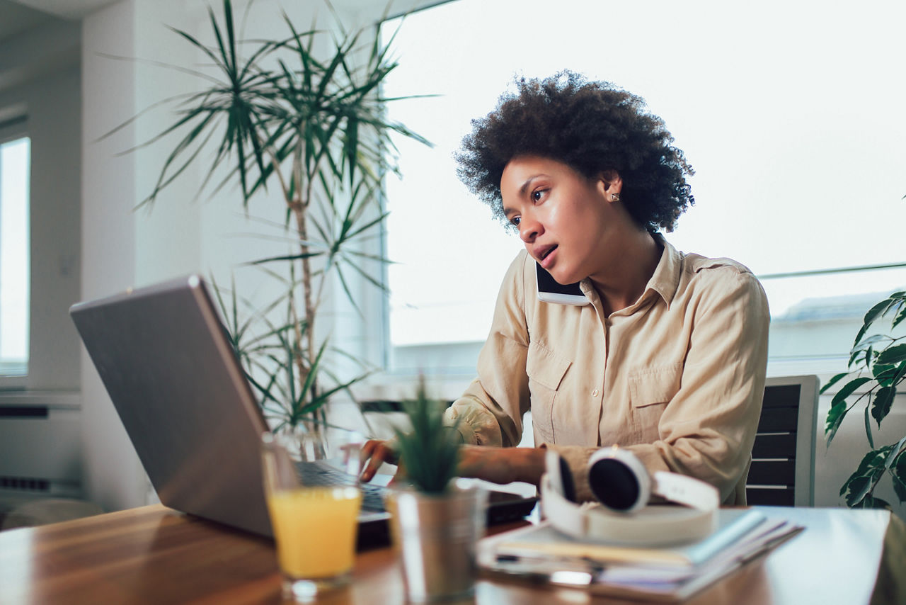 Smiling young African female entrepreneur sitting at a desk in her home office working online with a laptop