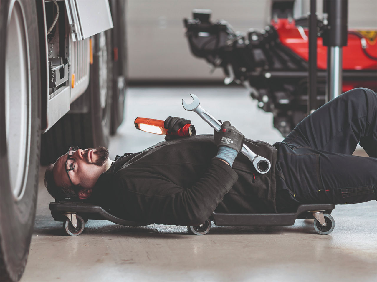 Service technician lying on a mechanic’s creeper beneath a truck, holding a wrench and inspection light during vehicle maintenance.