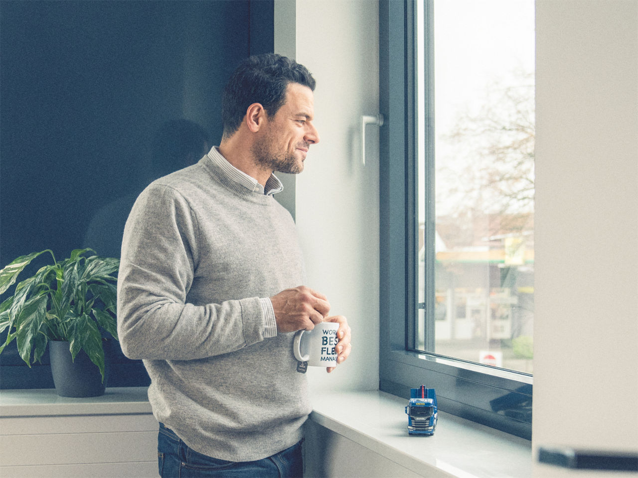 Un employé de bureau se tient debout près d’une fenêtre, tenant une tasse à café, regardant dehors, avec un modèle réduit de camion posé sur le rebord de la fenêtre.