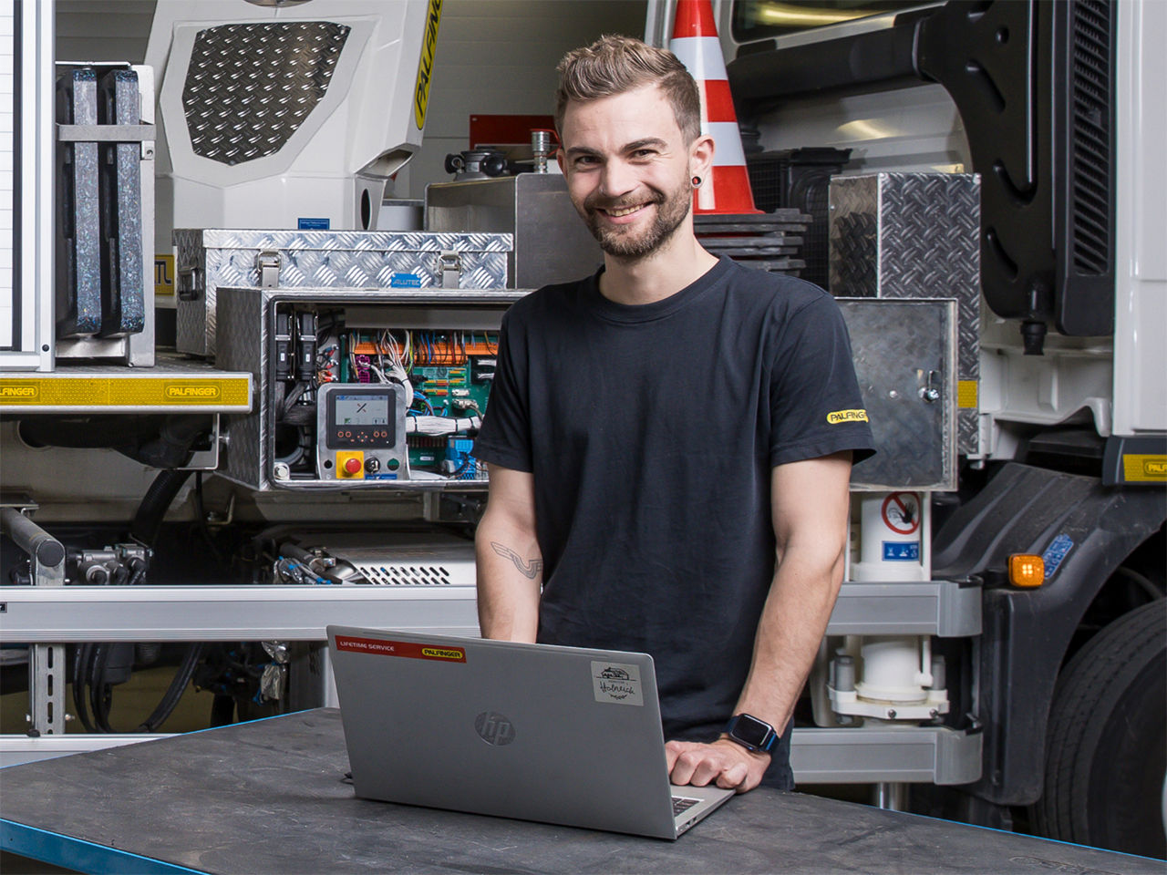 Smiling technician standing at a workbench with a laptop, in front of an open truck equipment compartment and control systems.