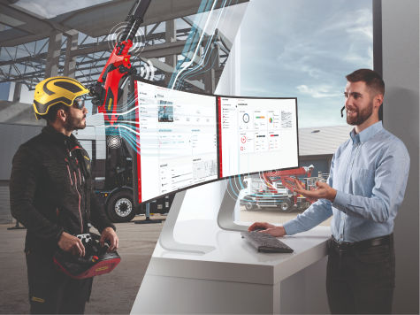 Un technicien industriel portant un casque de sécurité examine les données numériques de la flotte sur un grand écran tout en parlant avec un collègue à l’intérieur d’une installation de production.