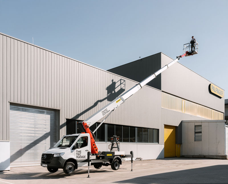 PALFINGER Hubarbeitsbühne PT 25 TJ aus der TEC-Reihe mit voll ausgefahrenem Ausleger und einem Bediener im Arbeitskorb vor einem PALFINGER-Werk.