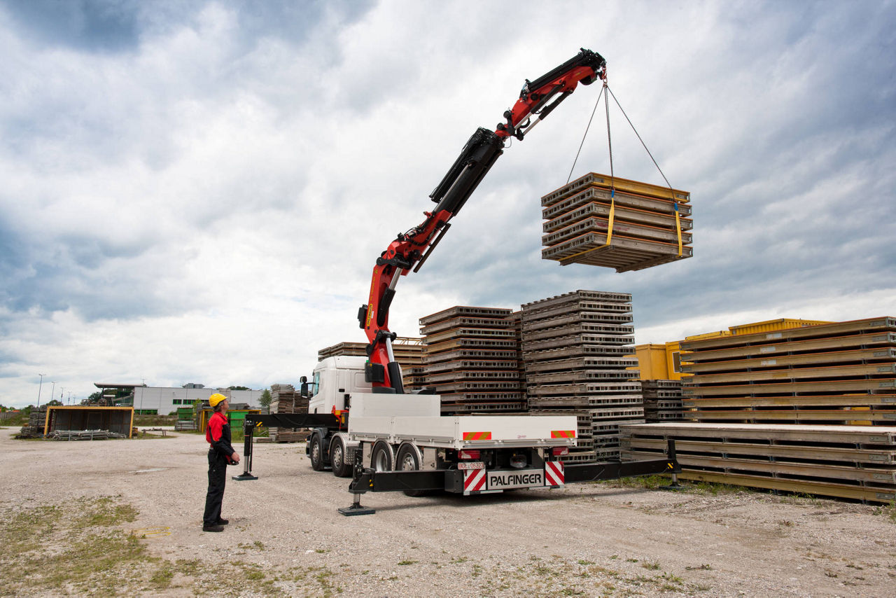 Truck-mounted crane lifting a bundled stack of wooden pallets at an industrial storage yard, guided by a ground operator.