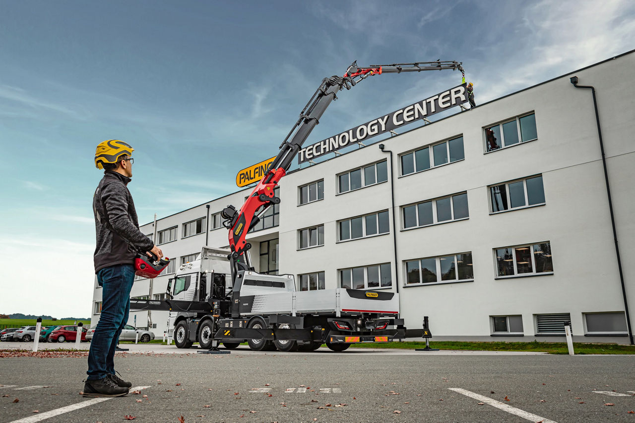 Operator using a remote control to position a truck-mounted crane in front of a building labeled “Technology Center.”