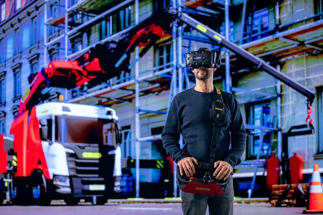 Operator wearing a VR headset and controllers in front of a PALFINGER truck-mounted crane at a job site.