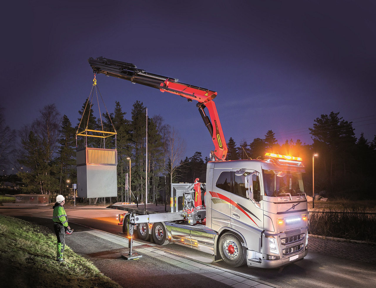Truck-mounted crane installing a large utility container at night, illuminated by vehicle work lights and guided by a ground worker.