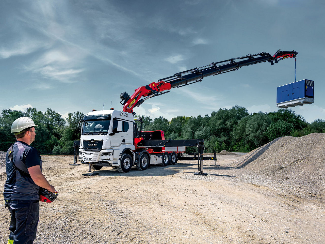 Truck-mounted knuckle boom crane lifting a blue industrial unit over a gravel site while an operator controls the crane remotely.