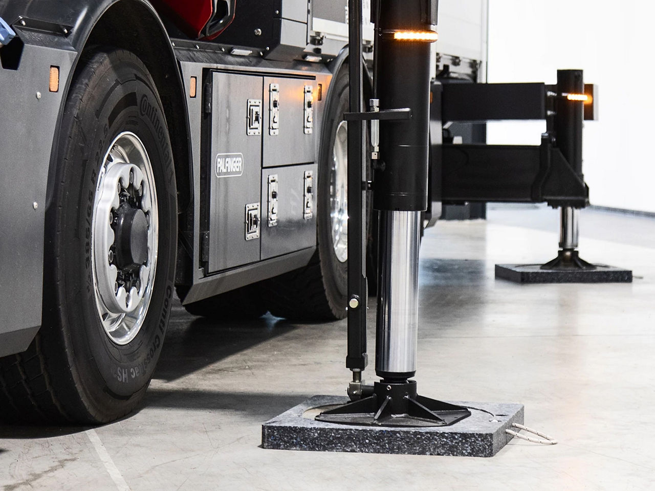 Close-up of a PALFINGER truck stabilizer leg deployed on a workshop floor, showing the wheel, hydraulic support arm, and base plate with sharp focus on mechanical details and materials.