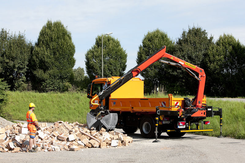 A yellow truck equipped with an orange Palfinger loader crane and a hydraulic grapple attachment picking up rubble and demolition debris at an outdoor work site, with a worker in orange high-visibility clothing and a yellow hard hat standing nearby.