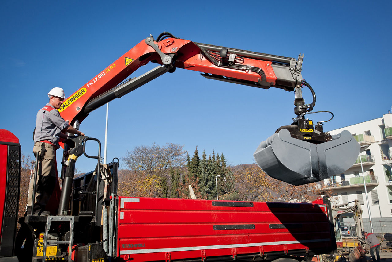 A worker in a hard hat operating a red Palfinger PK 17.001 SLD3 loader crane mounted on a truck, with a hydraulic grab attachment extended over a construction site on a clear sunny day.