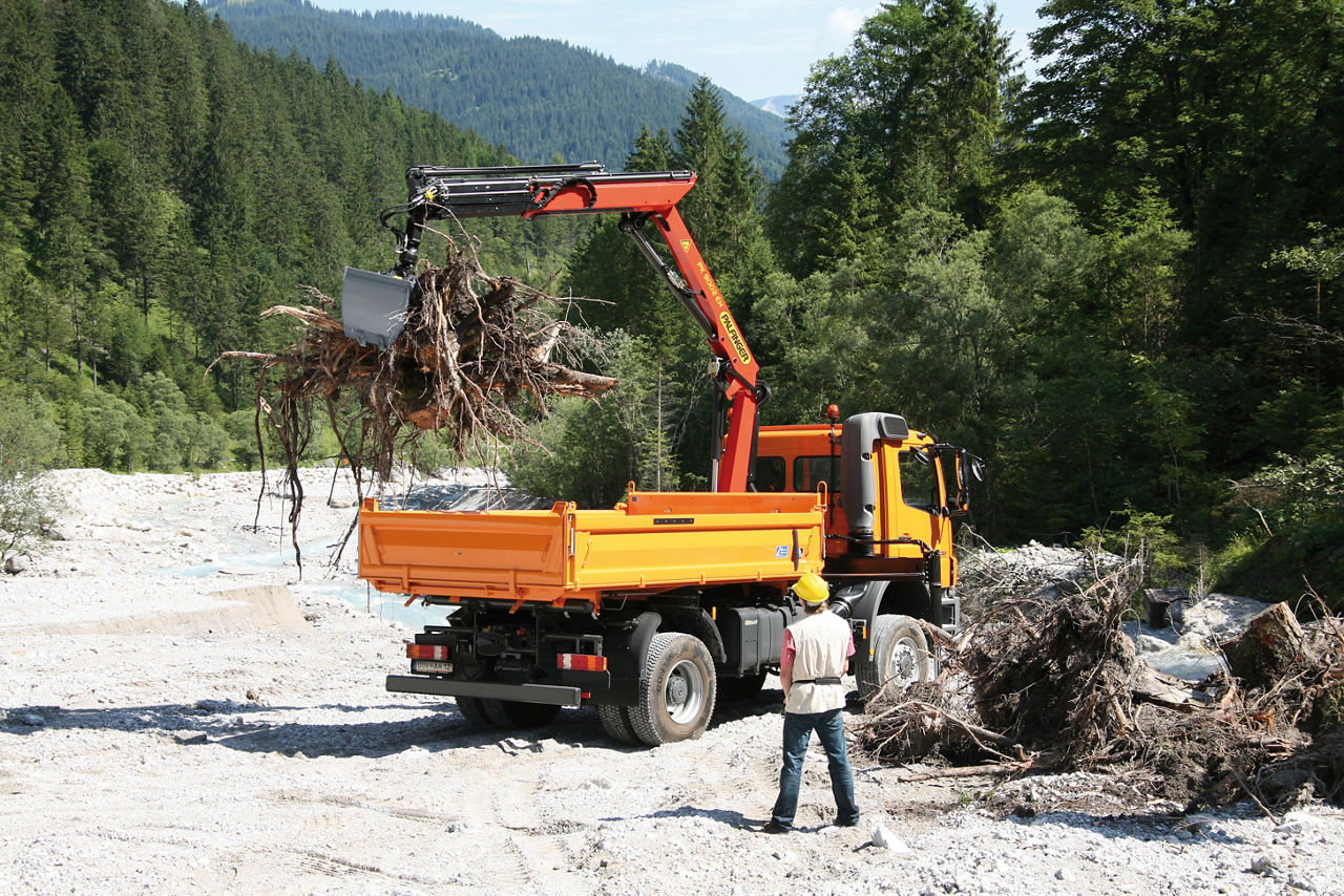An orange dump truck equipped with a Palfinger loader crane and grab attachment lifting uprooted tree debris at a river clearing or flood cleanup site in a mountainous forested landscape, with a worker standing nearby observing the operation.