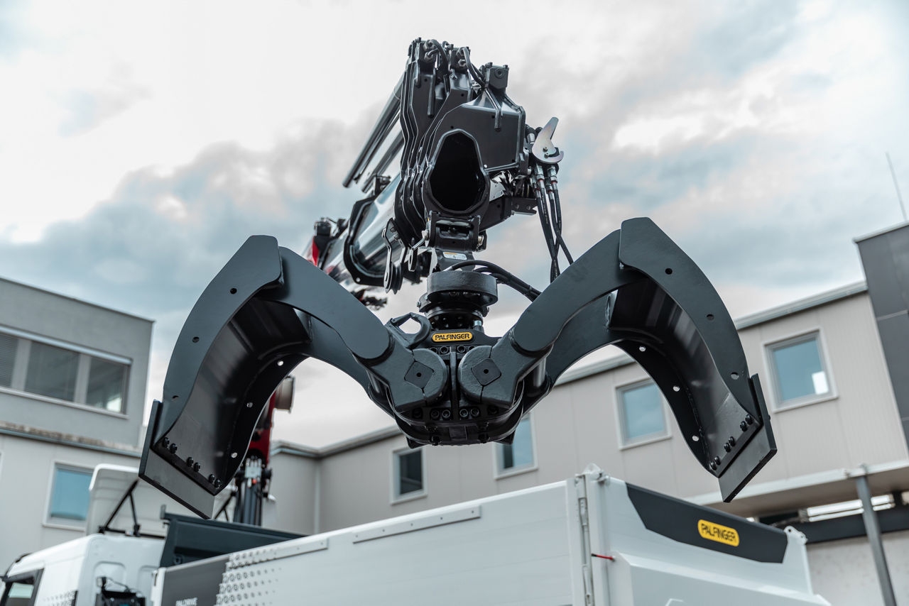 Close-up low-angle view of a PALFINGER hydraulic grapple attachment mounted on a loader crane above a truck, with open claws spread wide against a cloudy sky and a building in the background.