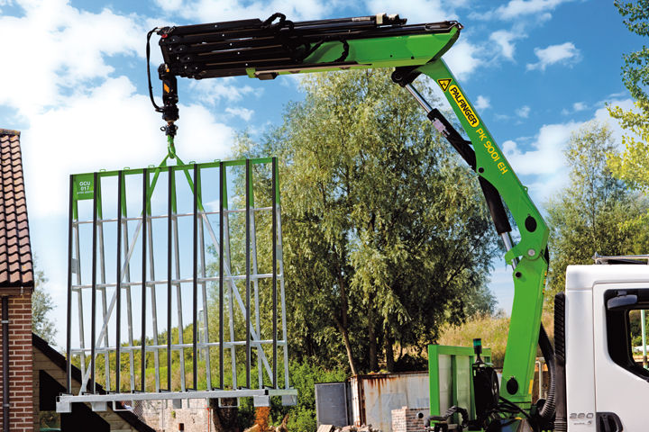 Green truck-mounted loader crane lifting a large steel frame structure at a residential construction site, with trees and a brick building visible in the background.