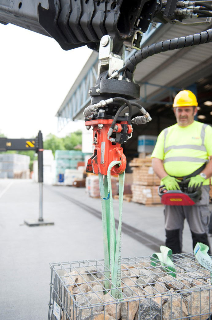 A loader crane hook with a red swivel connector and green lifting slings being used to lift a wire mesh gabion filled with stones, with a worker in a yellow hard hat and high-visibility vest operating a remote control in the background.