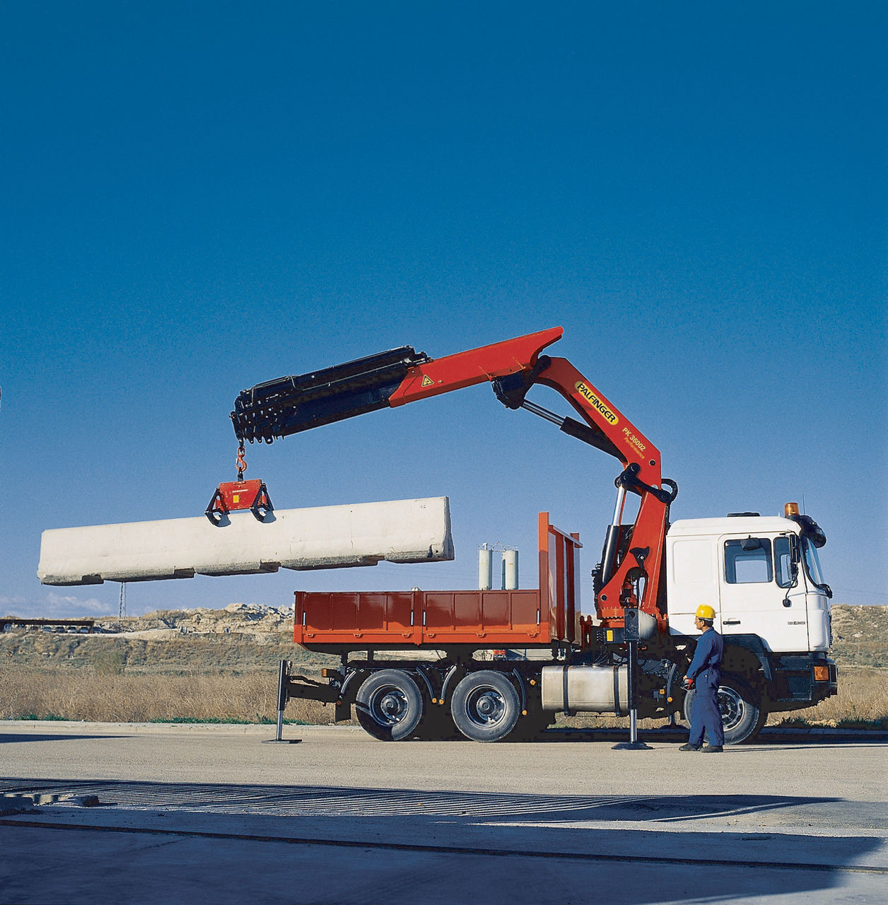 A red PALFINGER loader crane mounted on a white flatbed truck lifting a large precast concrete beam using a vacuum lifting attachment, with a worker in a yellow hard hat and blue overalls supervising the operation against a clear blue sky.