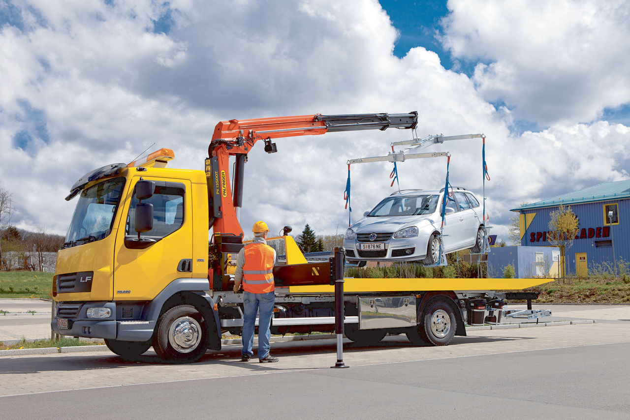 A yellow DAF LF flatbed recovery truck equipped with an orange PALFINGER loader crane lifting a silver Volkswagen passenger car using blue lifting slings, with a worker in an orange high-visibility vest and yellow hard hat operating the crane by remote control.