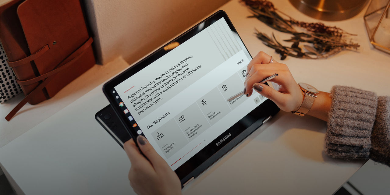 Person using a tablet with a stylus to navigate the PALFINGER website, showing business segments and a corporate mission statement on a clean desk setup.