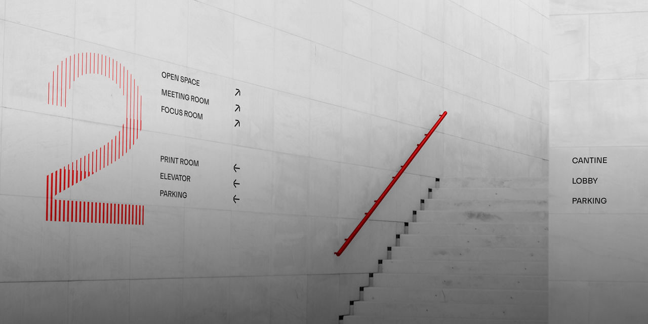 Minimalistisches Treppenhaus im Innenbereich mit hellgrauen Betonwänden, einem markanten roten Handlauf und Wegweisergrafiken, darunter eine große rote Zahl „2“ und Wegweiser zu Büroräumen, Lobby, Kantine und Parkplatz.