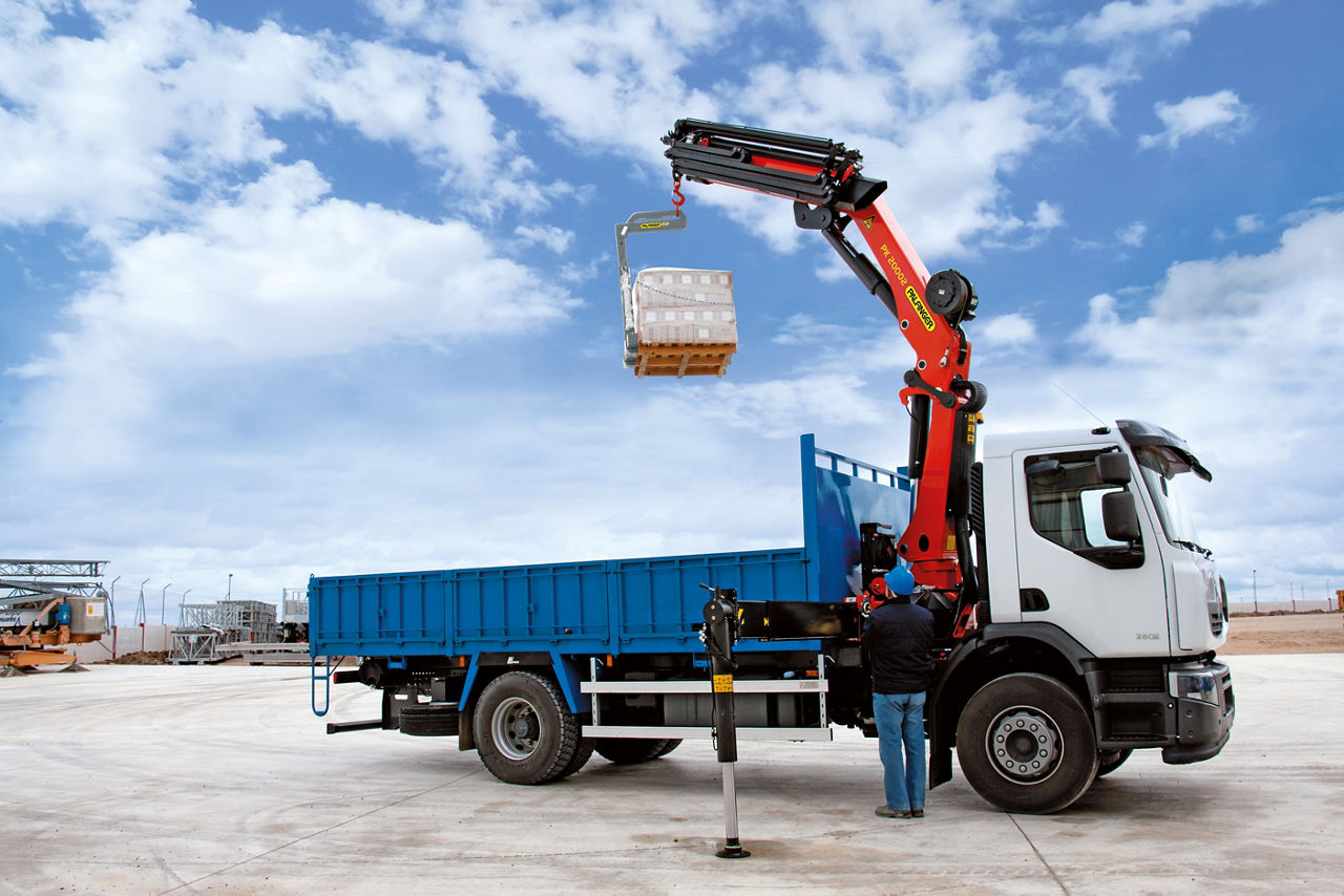A white truck with a blue flatbed body equipped with a red PALFINGER loader crane, lifting a pallet loaded with goods at an industrial site. A worker is standing next to the truck operating the crane under a partly cloudy sky.