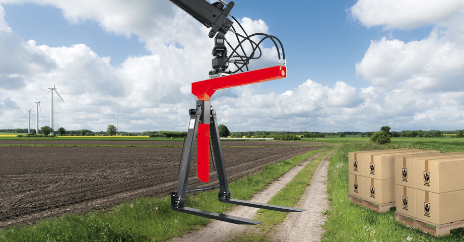 Hydraulic pallet fork attachment on a loader crane, used outdoors in an agricultural setting with stacked boxes on a pallet nearby.