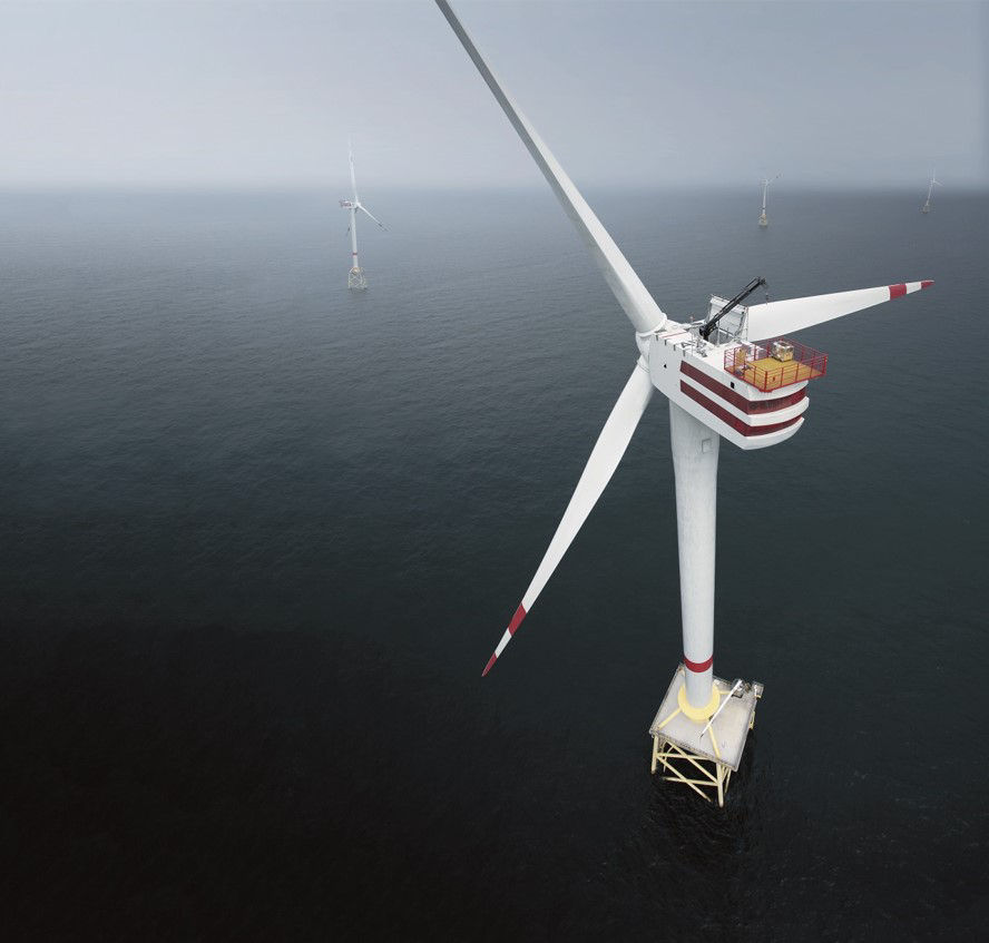 Aerial view of an offshore wind turbine rising from the sea, with long white blades extending over calm water and additional turbines visible in the distance, illustrating renewable energy generation at sea.