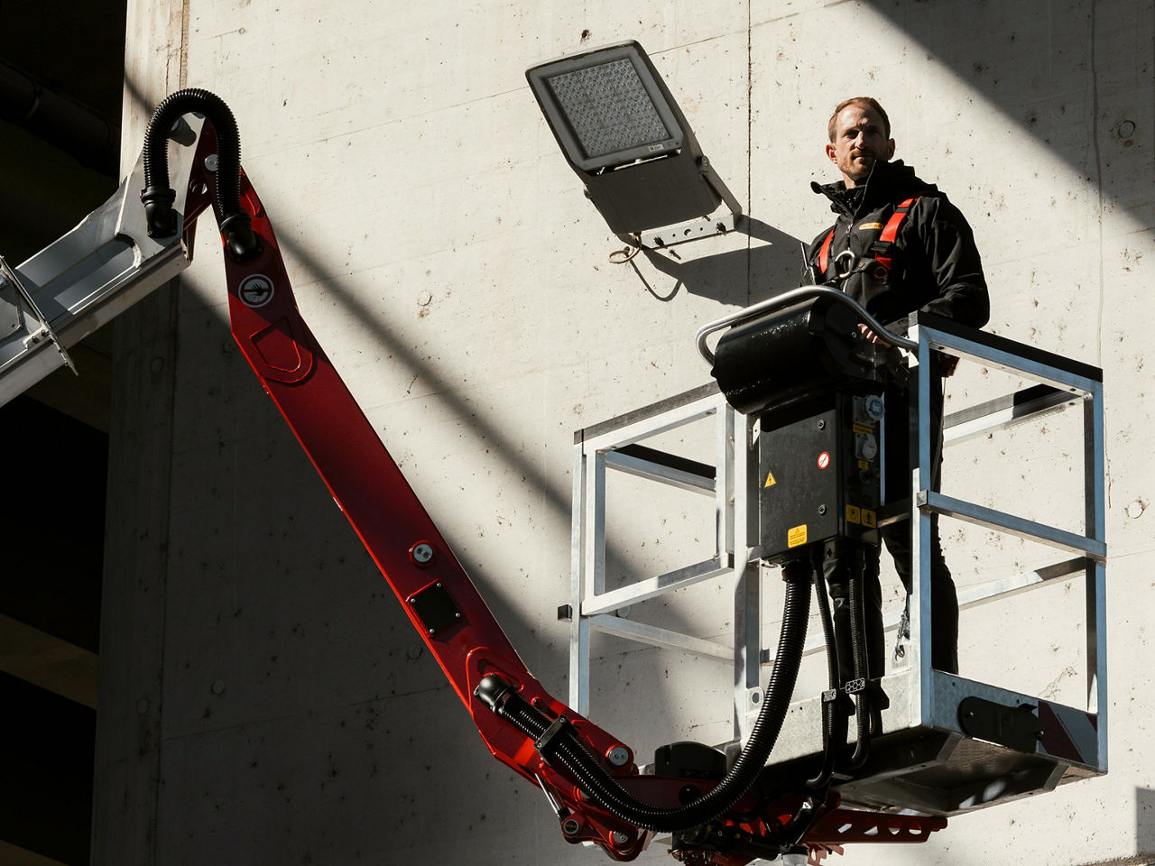 Operator standing in a lifting platform attached to a red crane arm, photographed with strong light and shadow to emphasize height and structure.