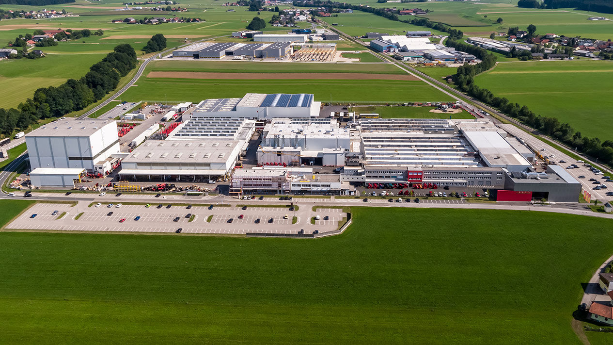 Aerial view of a large PALFINGER manufacturing facility surrounded by green fields and a parking area, showcasing industrial infrastructure in a rural setting.