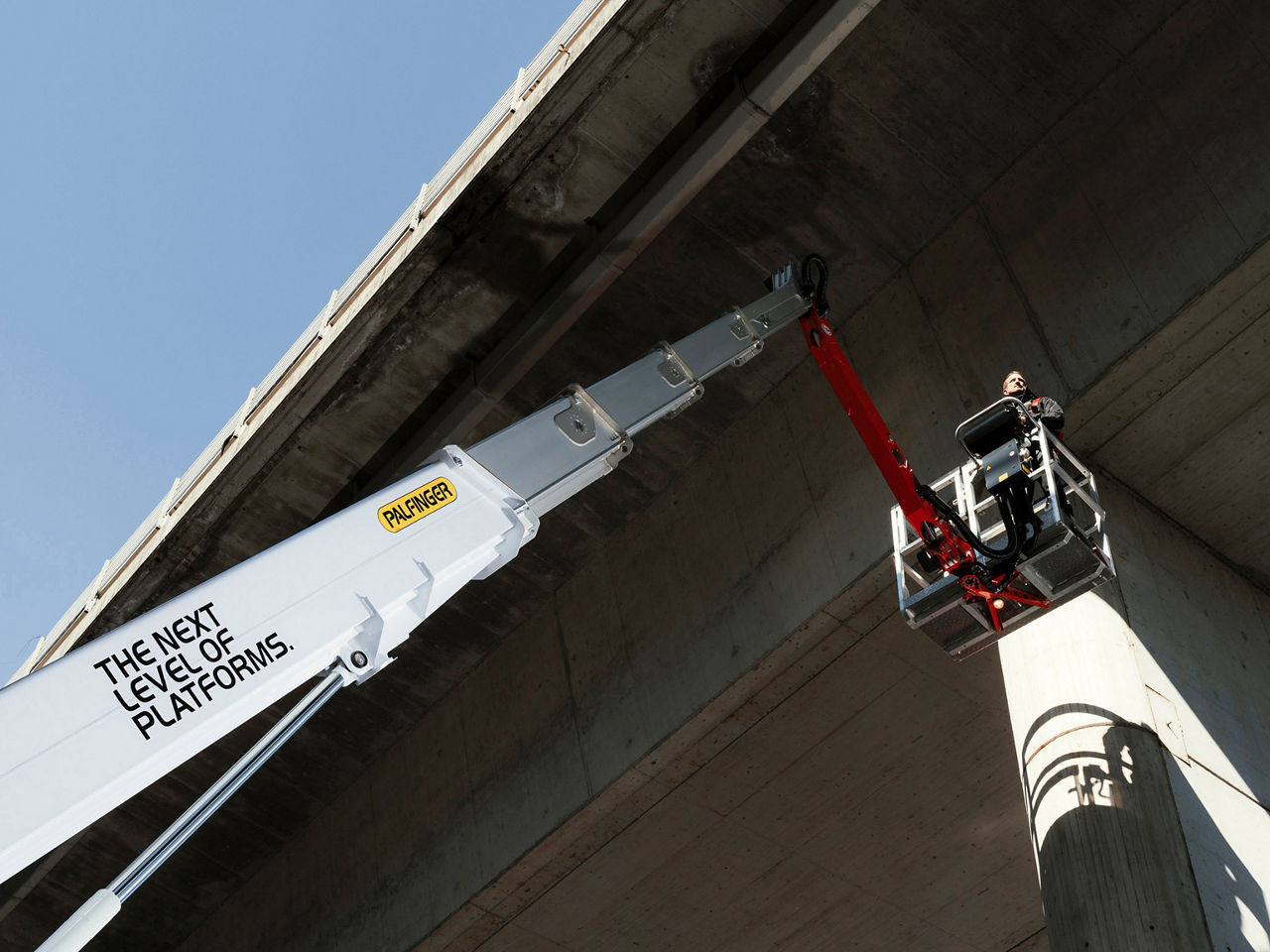 A lifting platform mounted on a crane operates beneath a concrete bridge, showing real-world use, human presence, and scale in an industrial environment.
