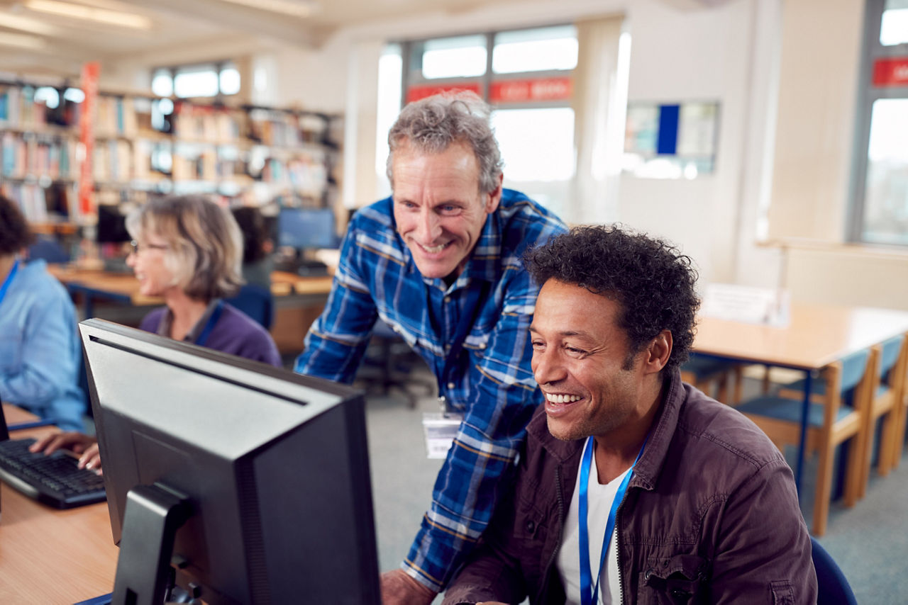 Teacher With Group Of Mature Adult Students In Class Working At Computers In College Library