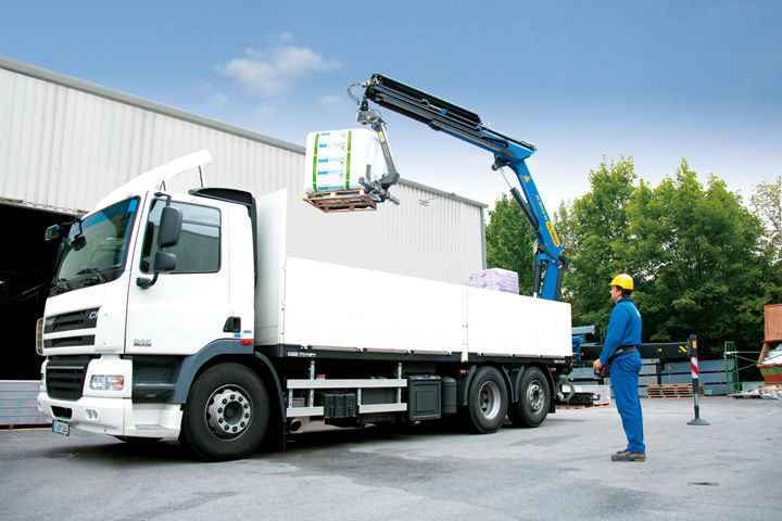 A white flatbed truck with a blue Palfinger loader crane uses a rotating fork attachment to lift a palletized load, guided by a worker in blue coveralls and a yellow safety helmet at an industrial site.