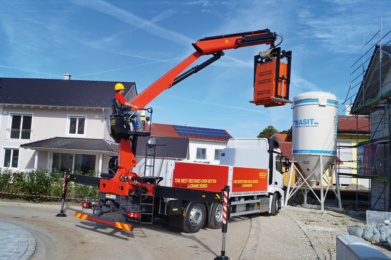 A red Palfinger loader crane mounted on a truck, equipped with a grabbing attachment (tongs/gripper), lifting a load at a residential construction site on a sunny day.