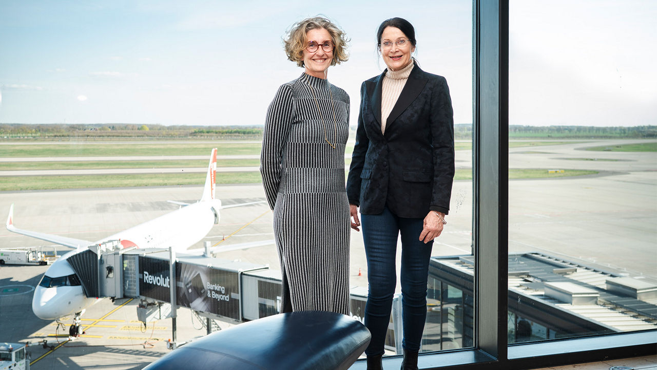 Zwei Frauen stehen vor einer großen Fensterfront am Flughafen. Im Hintergrund sind ein Flugzeug am Gate sowie das Rollfeld zu sehen