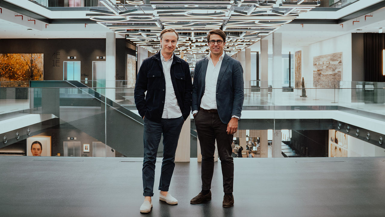 Two people standing on an upper-level walkway in a modern office building featuring glass railings, a multi-story atrium, and distinctive geometric ceiling lighting.
