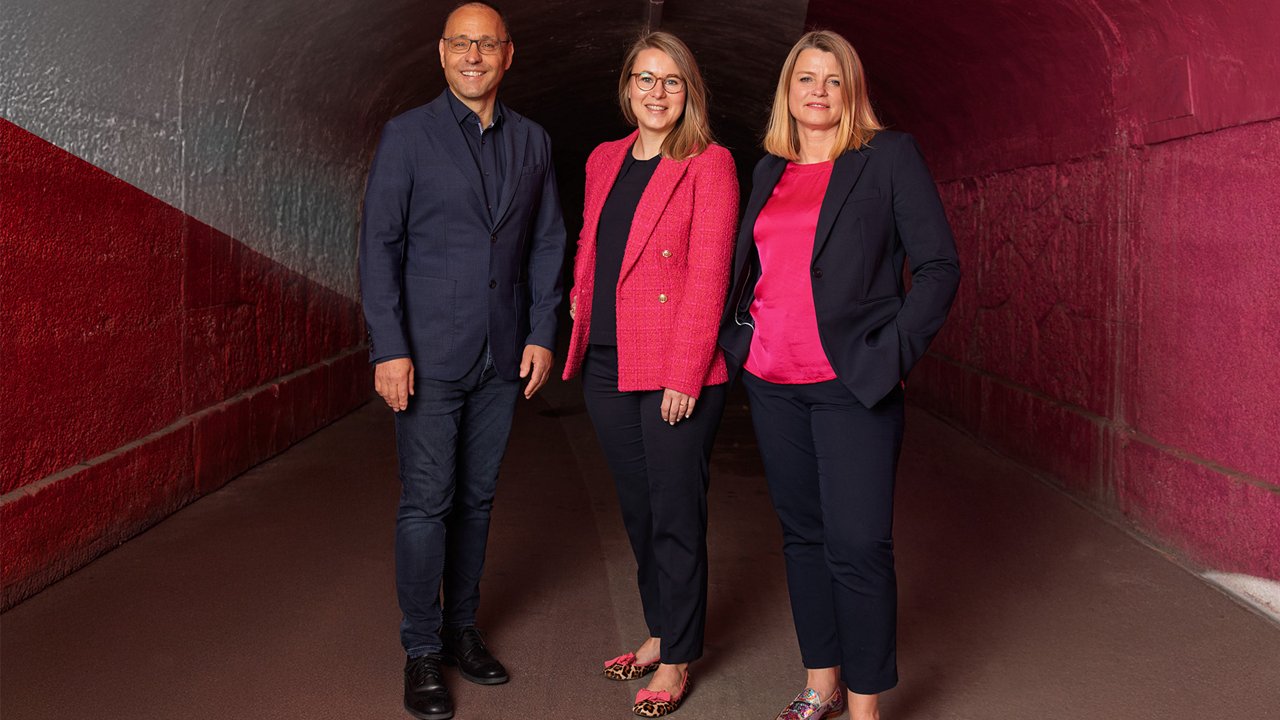 Hannes Haller, Isabell Decker und Claudia Raith stehen in einem rot-grau gestrichenen Tunnel nebeneinander, tragen Businesskleidung und blicken lächelnd in die Kamera..