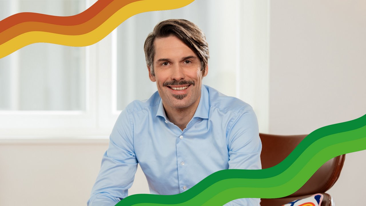 Jens Krahe, Managing Partner at Plan.Net Köln. Porträt eines freundlich lächelnden Mannes mit Schnurrbart in hellblauem Hemd, sitzend auf einem braunen Stuhl. Im Vordergrund verlaufen dekorative Wellenlinien in Rot-, Gelb- und Grüntönen..