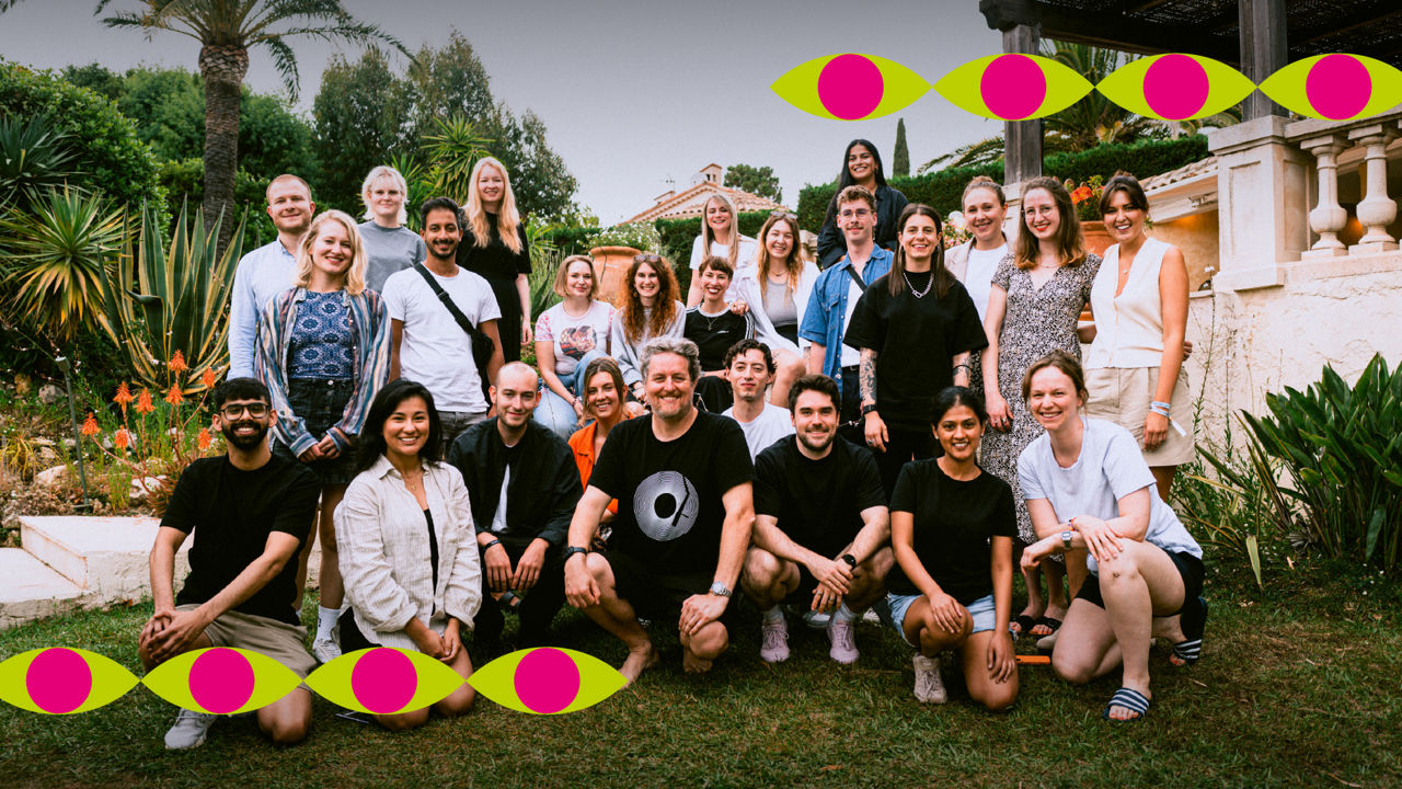 Group photo of around 25 people in a garden in front of a Mediterranean-style villa. The group is casually seated and standing among palm trees and flowers. Colorful eye-shaped graphic elements frame the top and bottom of the image.