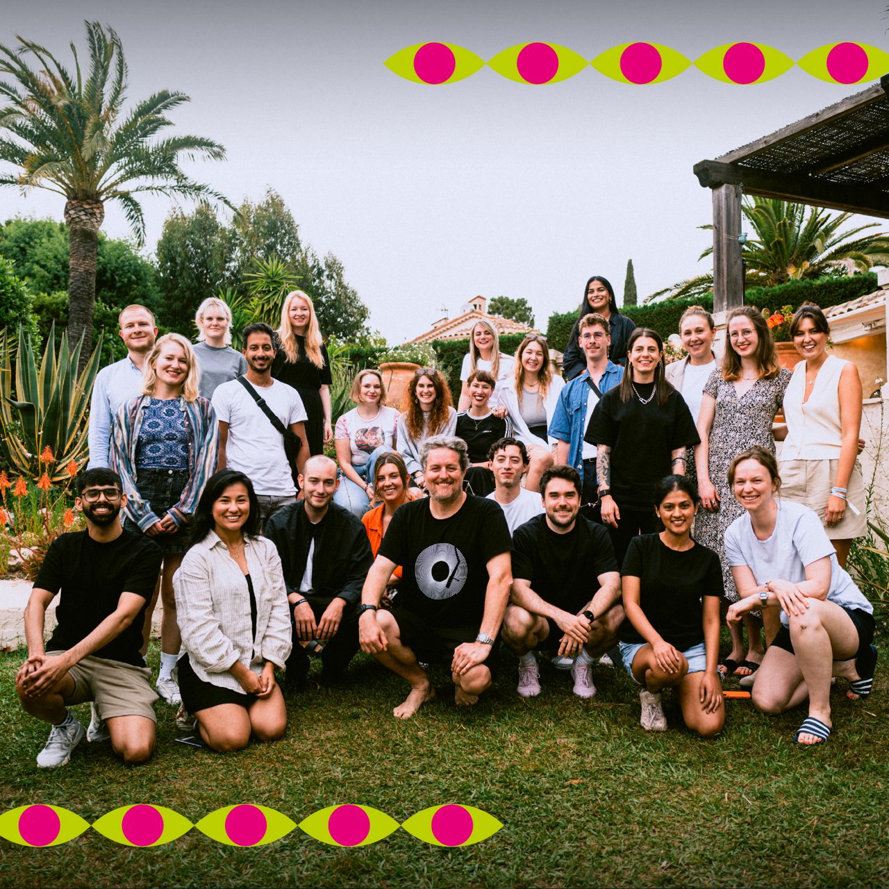 Gruppenfoto von etwa 25 Personen vor mediterraner Villa im Garten. Die Gruppe steht und sitzt entspannt, umgeben von Palmen und Blumen. Bunte grafische Augensymbole zieren den oberen und unteren Bildrand.