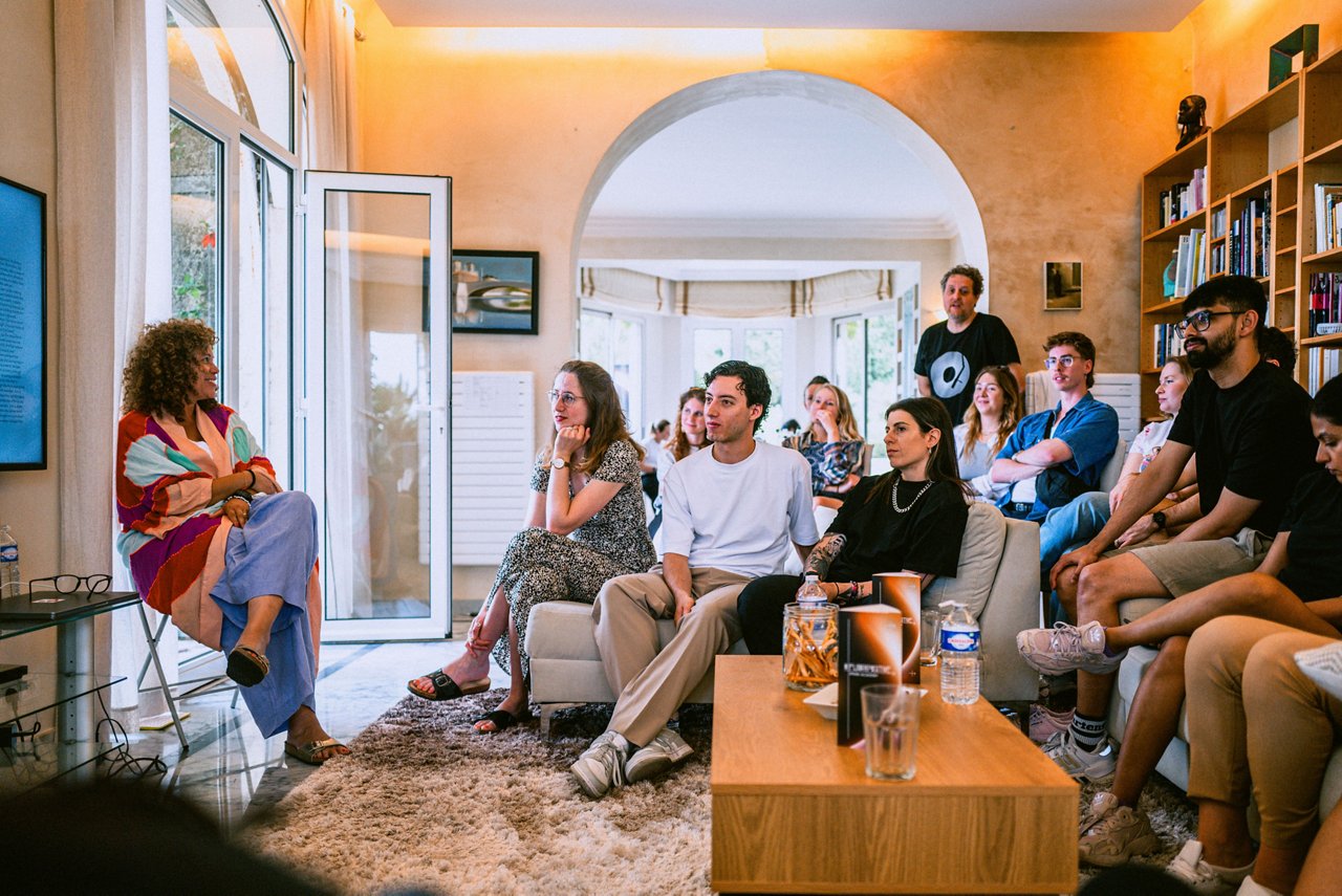 Spark Academy 2025 in Cannes: A woman in colorful clothing gives a talk to a seated group in a living room with bookshelves.