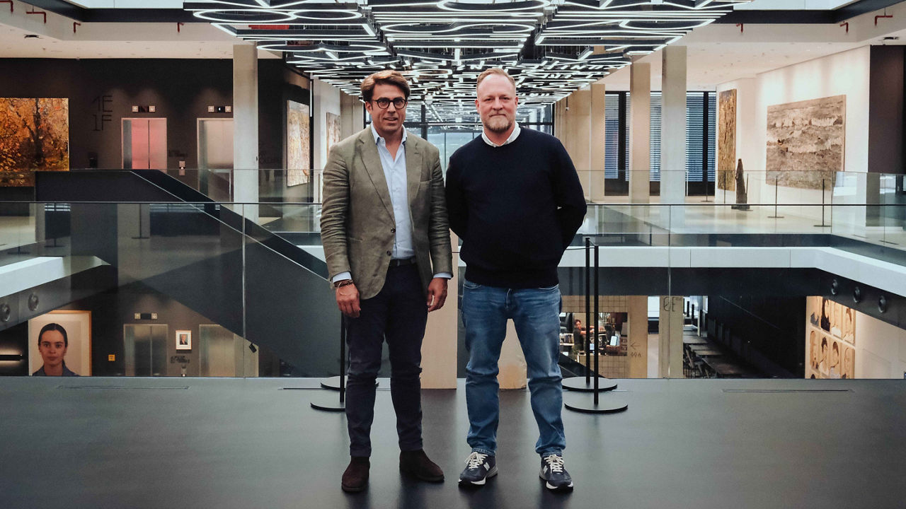 Two people are standing in a modern atrium featuring multiple levels, glass railings, and distinctive ceiling lighting. Large-scale artworks and open office spaces are visible in the background.