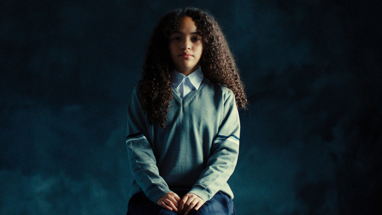 A girl with curly hair in a school uniform sits against a dark background, looking calmly at the camera.