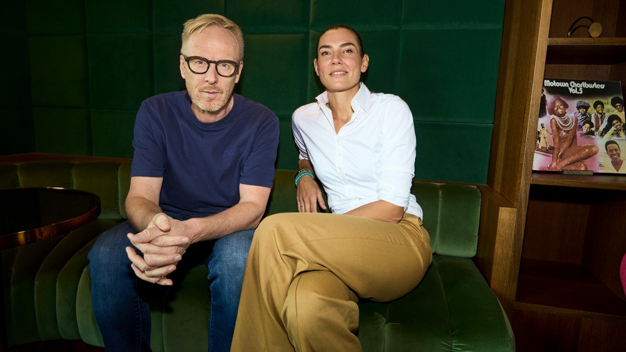 Saint Elmo's Group CCO Matthias Harbeck und CEO Kathrin Jesse. Ein Mann mit Brille und eine Frau in weißer Bluse sitzen nebeneinander auf einem grünen Sofa in einem Raum mit dunkler Wandverkleidung. Im Regal hinter ihnen steht eine Schallplatte mit Soul-Musik-Cover.