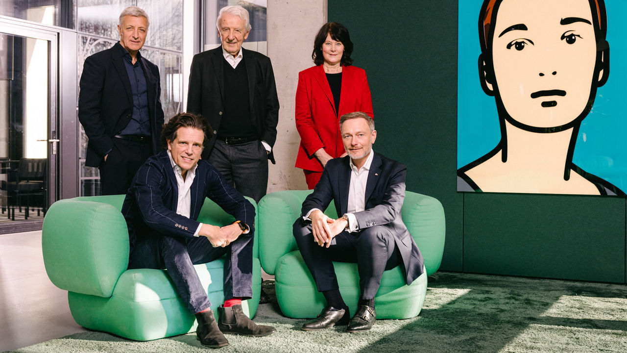 Five adults in business attire pose in a modern office: two are seated in green armchairs, while three stand behind them. On the wall to the right hangs a large, stylized portrait.