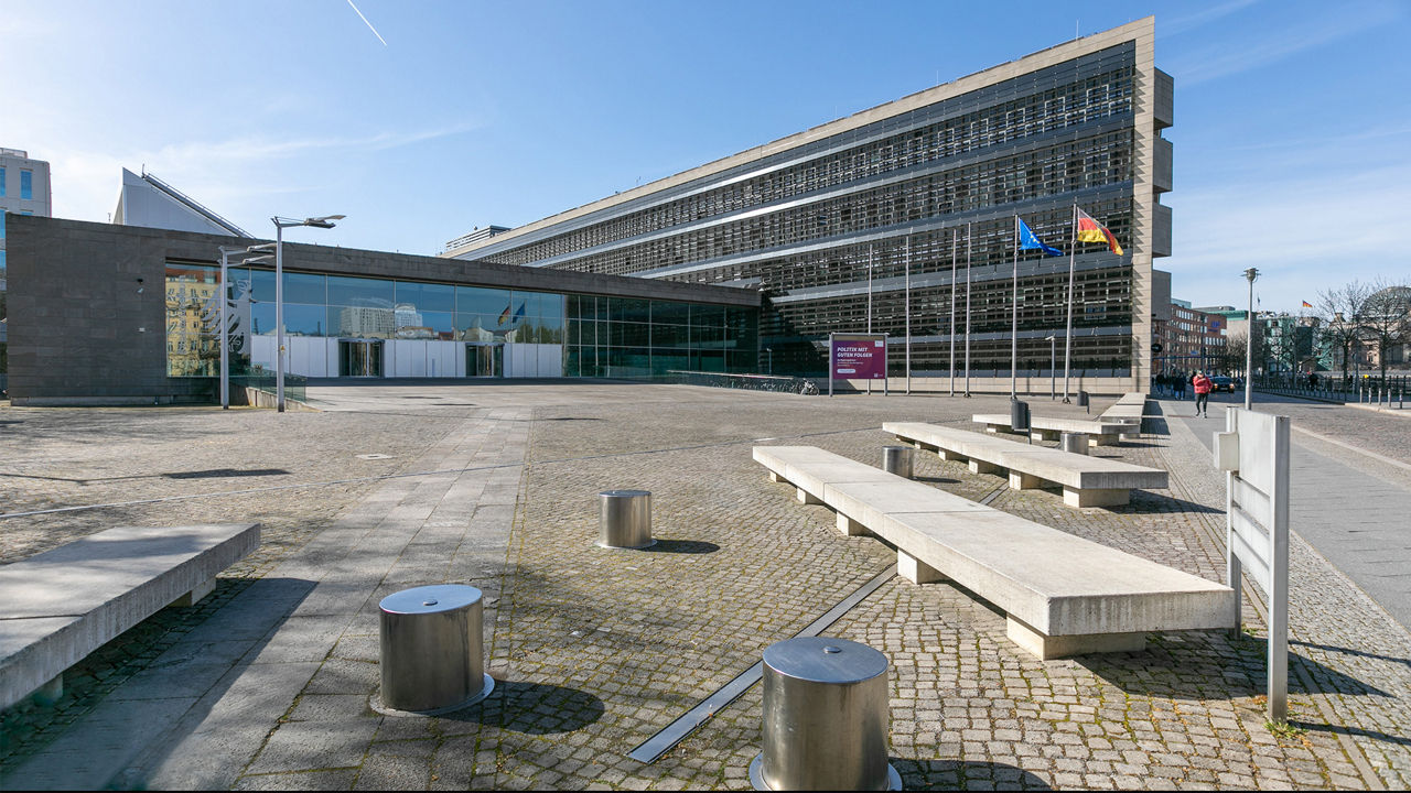Modernes Verwaltungsgebäude mit Glasfassade und Betonstruktur auf einem offenen, gepflasterten Platz bei klarem Himmel. Vor dem Gebäude wehen die Flaggen der Europäischen Union und Deutschlands. Im Hintergrund sind einige weitere Gebäude sichtbar, eine Person geht über den Platz.
