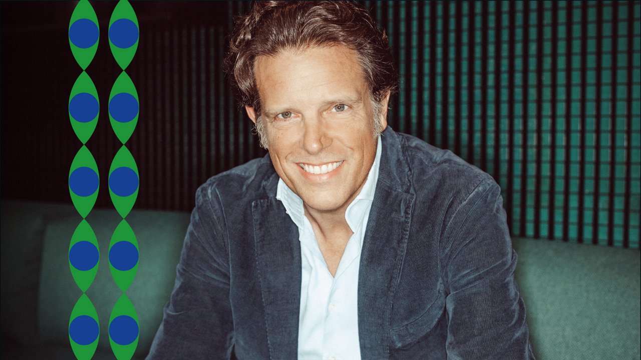 Portrait of Florian Haller, CEO of Serviceplan Group. He is sitting on a sofa, wearing a light blue shirt and a dark blue corduroy jacket. He is looking directly at the camera with a broad, open smile showing his teeth. Vertical dark green structures are visible in the background.