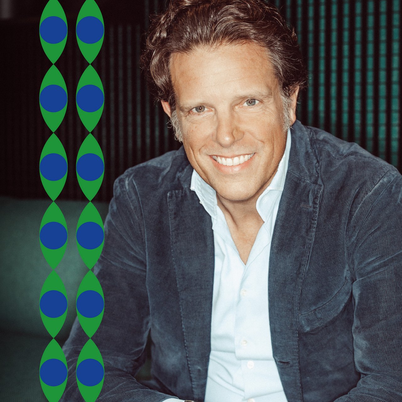 Portrait of Florian Haller, CEO of Serviceplan Group. He is sitting on a sofa, wearing a light blue shirt and a dark blue corduroy jacket. He is looking directly at the camera with a broad, open smile showing his teeth. Vertical dark green structures are visible in the background.