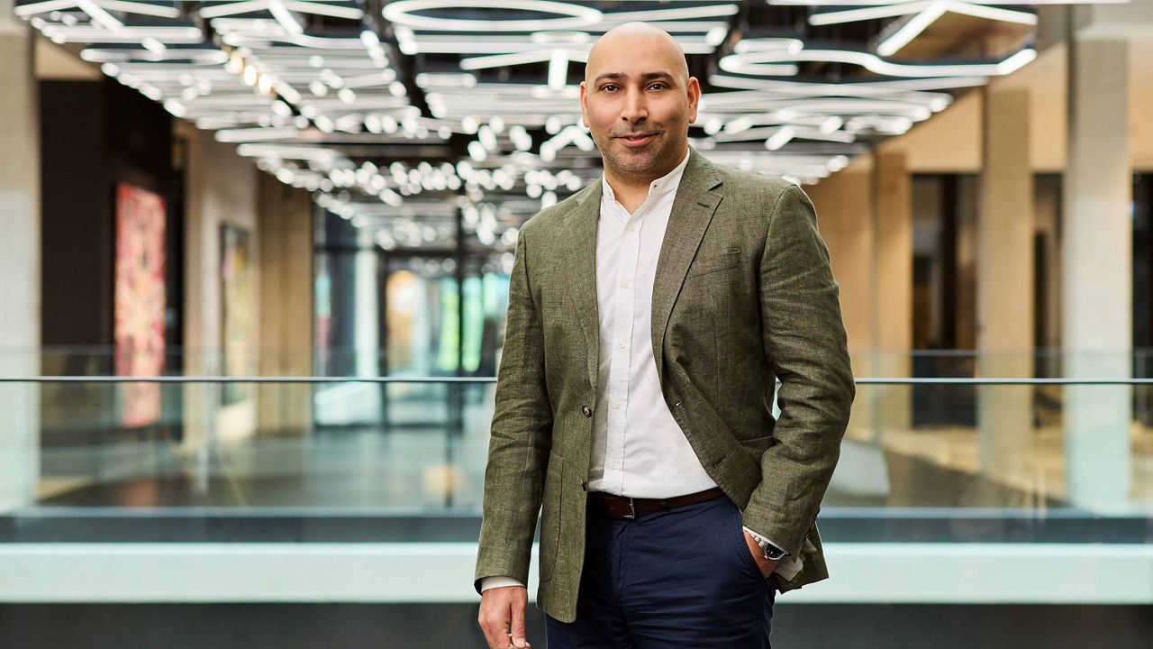 Person standing in a modern office building, wearing a light shirt, green blazer, and dark trousers, with geometric ceiling lights in the background.