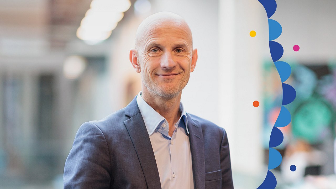 Portrait of John Wittesaele. He is standing in a modern office building, wearing a light blue shirt and a dark blue blazer. He is leaning slightly against a railing with one arm and looking at the camera with a subtle smile. The background is blurred, showing bright structures and colorful wall art.